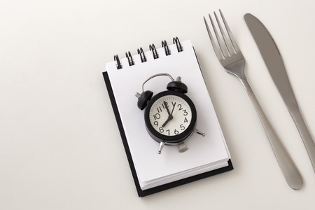 Clock on notepad with fork and knife for intermittent fasting and weight loss plan concept on white backgroundの写真素材