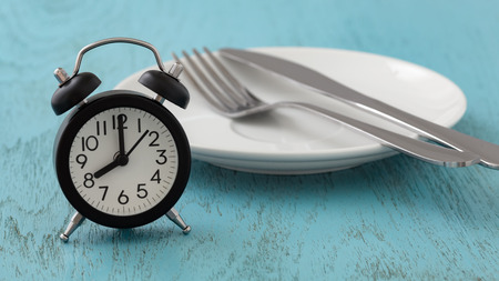 Intermittent fasting concept with clock, white plate, fork and knife on blue table