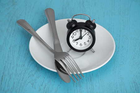 Intermittent fasting concept with clock on white plate, fork and knife on blue tableの写真素材