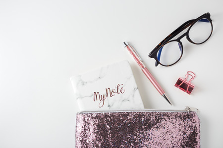 Glitter bag with pen, planner notebook and eyeglasses, pink gold accessories on white deskの写真素材