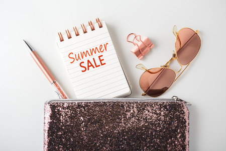 Summer sale word on notepad with glitter bag, pen and sunglasses, pink gold accessories on white deskの写真素材