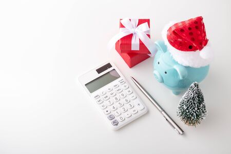 Piggy bank wearing christmas hat with red gift box, calculator and pen on white table, with copy spaceの写真素材