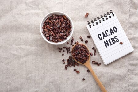 Cacao nibs in white bowl and wooden spoon with handwritten note on table, top view with spaceの写真素材
