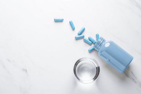 Blue pill capsules spilling out of bottle and glass of water on white background, top view, copy...