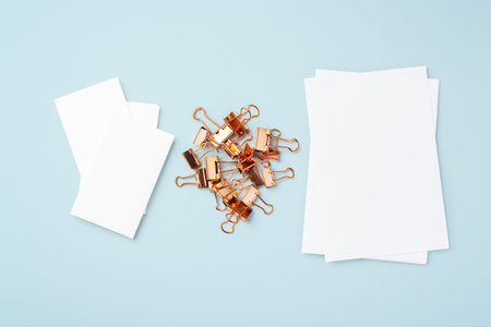 Heap of gold binder clips and memo note paper on light blue background, top view