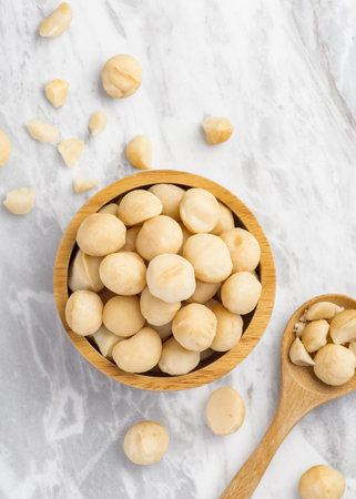 Macadamia nuts in wooden bowl with spoon and cracked macadamia on marble table background, top viewの写真素材