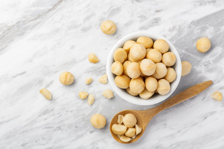Macadamia nuts in white bowl with wooden spoon and cracked macadamia on marble table background, top view, copy spaceの写真素材