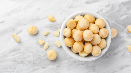 Macadamia nuts in white bowl with cracked macadamia on marble kitchen countertop background,...