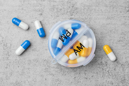 Daily pill box organizer with AM and PM compartments with blue and yellow capsules on a grey concrete background, top view