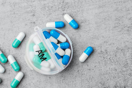 Capsule pills spilling with open daily pill organizer on a grey concrete background, copy space