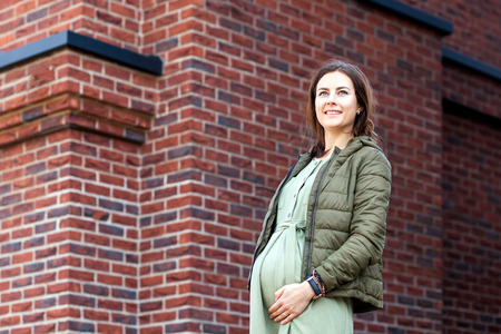 Modern pregnant woman in a light jacket and casual dress posing against a brick house backgroundの写真素材