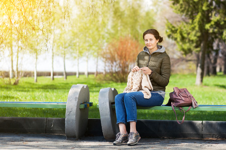 On a summer day in the city park A young brunette woman knits a scarf with knitting needles and sits on a benchの写真素材