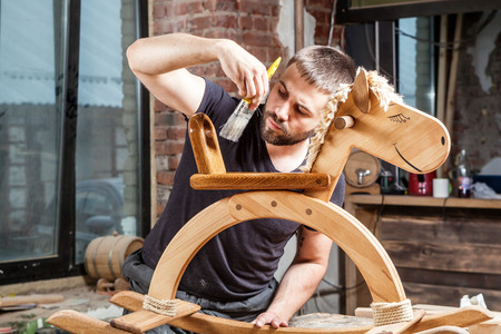 Young man builder carpenter paints children's wooden toy rocking chair in the form of a horseの写真素材