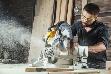 Close-up as a young male construction worker carpenter saws a circular saw blade, sawdust fly in the...