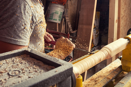 A young male construction worker carpenter processes a wood d on a lathe in the workshop, close-up on his hands and around a lot of wooden sawdust, in the background a woodenの写真素材