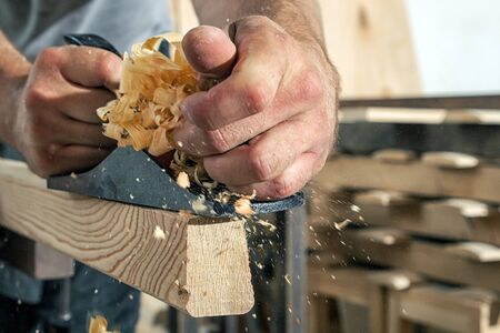 A close-up as a Young man is treating a wooden bar with a black jack plane in the workshop, in the background a lot of wooden boardsの写真素材