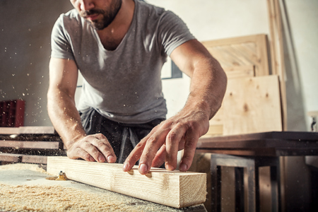 A young male carpenter builder in a gray T-shirt and working overall equals a wooden bar with a milling machine in the workshop, in the background wooden boardsの写真素材