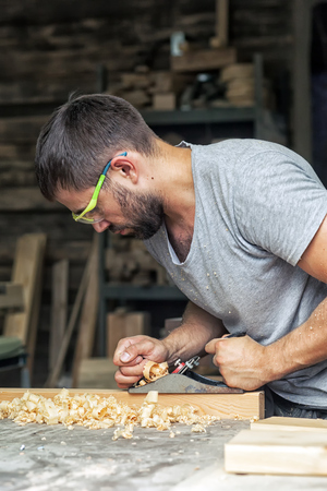 A young man with a beard and in sunglasses by profession, the carpenter holds a black jack plane in his hands and equals a wooden board, in the background a lot of wooden boards and equipmentの写真素材