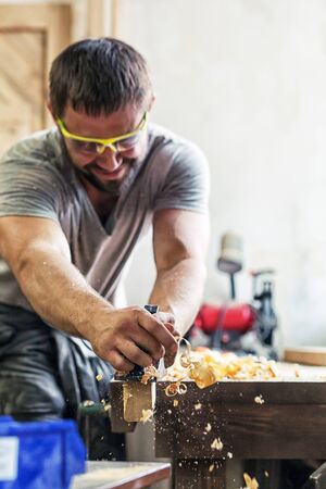 A young man with dark hair and wearing goggles is holding a black plane in his hands and is processing a wooden board in his hand flying wood sawdust in the workshopの写真素材