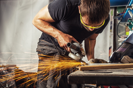 A young man is a brunette welder wearing a black T-shirt and safety goggles grinding metal with a angle grinder in the workshop, sparks flying in the sidesの写真素材