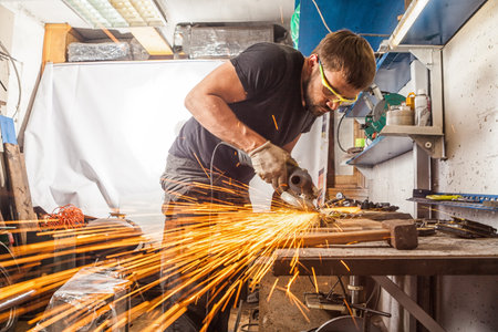 A young man welder in a black T-shirt, goggles and construction gloves grinder metal an angle...