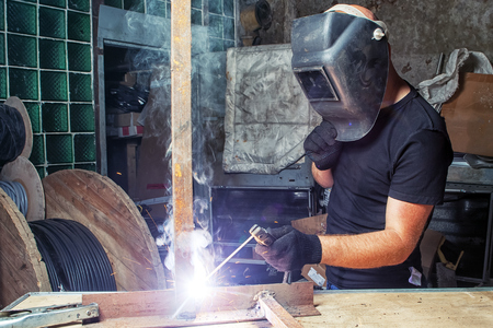 A bald, strong man in a black T-shirt welds a metal welding machine in the production hall, a green glass mill on the background,  there are a lot of tools on the table for workの写真素材