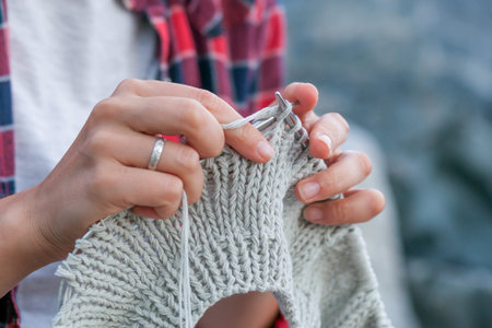 Close-up Young woman checkered red shirt and white t-shirt knits with knitting needles gray sweater...