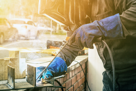 Man welder in welding mask, building uniform and blue protective gloves brews metal welding machine on street construction, in the background an old brick building with window and toolsの写真素材