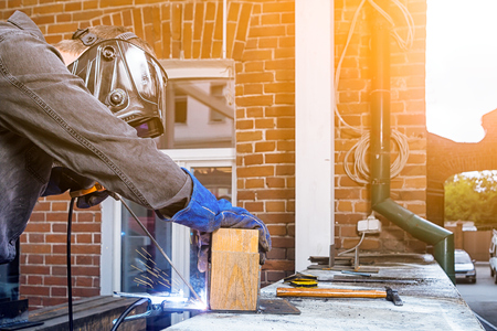 Man welder in welding mask, building uniform and blue protective gloves brews metal welding machine on street construction, in the background an old brick building with window and toolsの写真素材