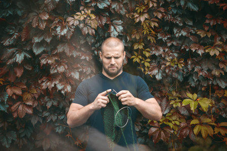 A young handsome man stands on a background of a green wall of real and grapes and knits a green sweater with the knitting needles of their natural woolen threads. A man learn to knit  needles sweaterの写真素材