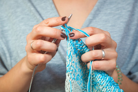 A close-up of a young married woman in a gray cardigan knits with knitting needles from natural woolen threads a white and blue sweater. Close-up of female hands with brown manicure and short nailsの写真素材