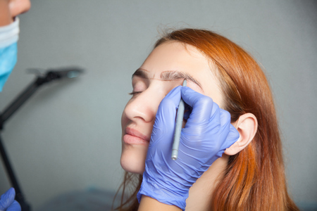 The master on the eyebrows draws the shape of the grafted eyebrow shape in a blue medical disposable gloves red-haired beautiful woman on a gray backgroundの写真素材