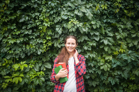 Young dark-haired woman in a red shirt in a box stands smiling and holding a green and red book on the background of a living wall of natural green grapesの写真素材
