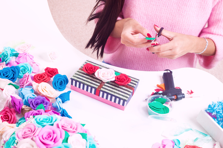 A young woman makes a striped casket and cuts on it artificial flowers - roses from foam different colors for decorating weddings, interiors , a knife on the table, a glue gun, boxes and petalsの写真素材