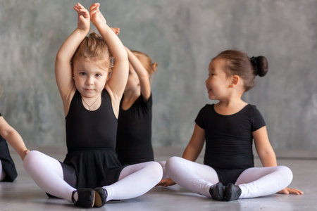 A blonde girl and a small Asian girl in black swimsuits, white tights and pointe shoes sit on the pound and stretch before a ballet in a dark dance studioの写真素材