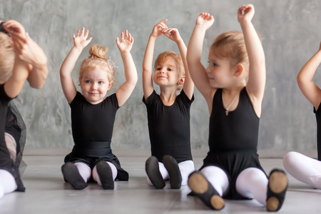 little girls ballerina in black dresses, belk tights and pointe shoes sit on the floor and hold hands over their head for warm-up before teaching ballet in a dancing dark studioの写真素材