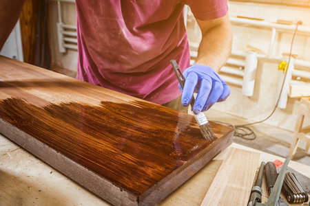 Close-up of a man with work clothes and protective gloves paints a wooden board with a protective paint brush in a light workshopの写真素材