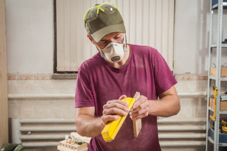 A man in a cap, safety mask, goggles and a T-shirt is sanding a wooden  in the workshop, side view,...