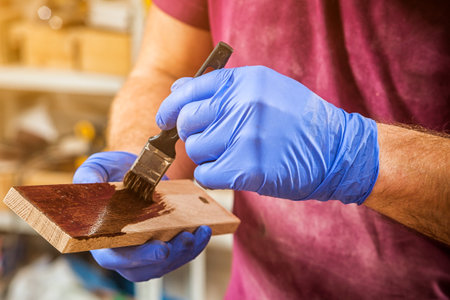 Close-up of a man in blue protective gloves paints a wooden board with a brown wooden brush with a brown paint for protection in workshopの写真素材