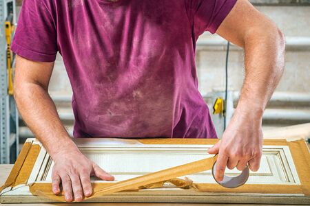 A working man in a green cap and t-shirt a wooden frame with a double tape to protect himself from paint in the workshop, in the background, tools and equipmentの写真素材