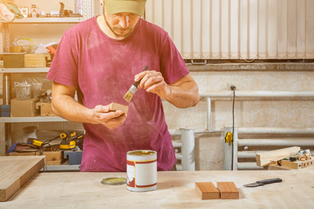 Close-up The man carpenter in working clothes paints a wooden block with a brown protective paint...