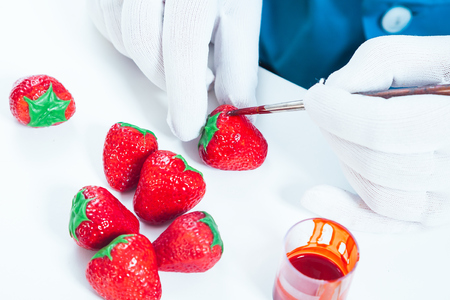 A woman confectioner with blue uniform and white sterile gloves paints a nophet in the form of a strawberry in red with edible paint on a white tableの写真素材