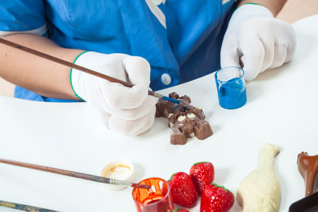 A woman confectioner with blue uniform and white sterile gloves colors a dog-shaped candy in golden color with edible paint on a white table for a New Year's gift, on the table there are other candiesの写真素材