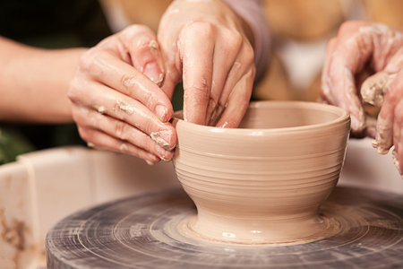 A close-up of a woman potter teaches a woman how to properly mold a bowl of brown clay on a potter's wheel in a bright workshop, side view. Training of modeling on a potter's wheelの写真素材