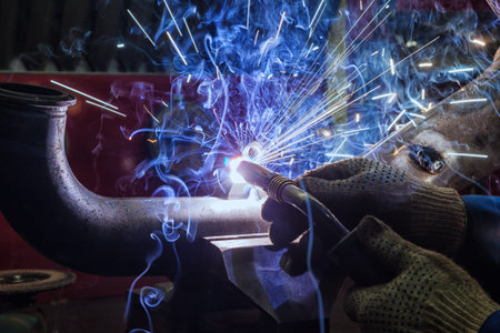 Close-up of a welder in protective gloves welds a metal pipe with a welding machine, blue sparks...