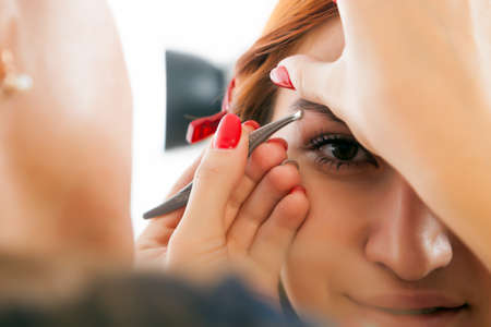 A young woman with a tassel plucks out her red-haired woman's eyebrows to achieve the ideal shape in a beauty salon, front viewの写真素材