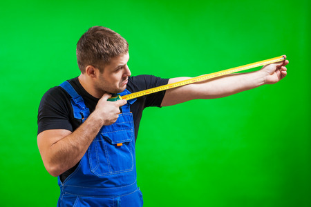 A dark-haired male builder in a black T-shirt and blue construction jumpsuit demonstrates and measures with his long yellow tape rule his hand on a green isolated backgroundの写真素材