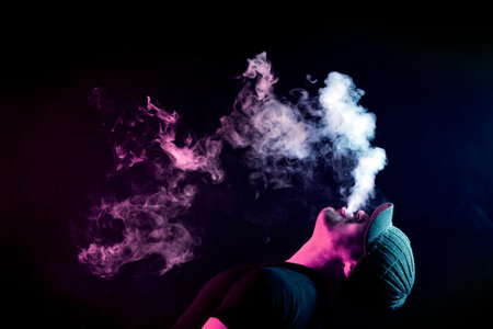 A young man exhales a cloud of colored smoke of red color on a black isolated color all the back is covered with yellow smoke. Harm to smoking on the example of a young muggerの写真素材