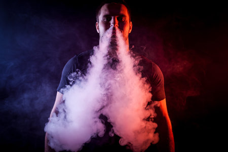 A young stylish male smoker in a black T-shirt exhales a large cloud of smoke from vape on a black...