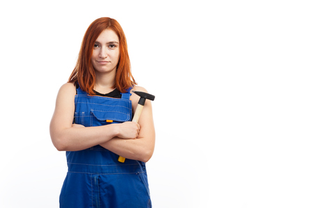 Young attractive woman in blue construction overalls smiling and holding a black wooden  hammer on white isolated backgroundの写真素材